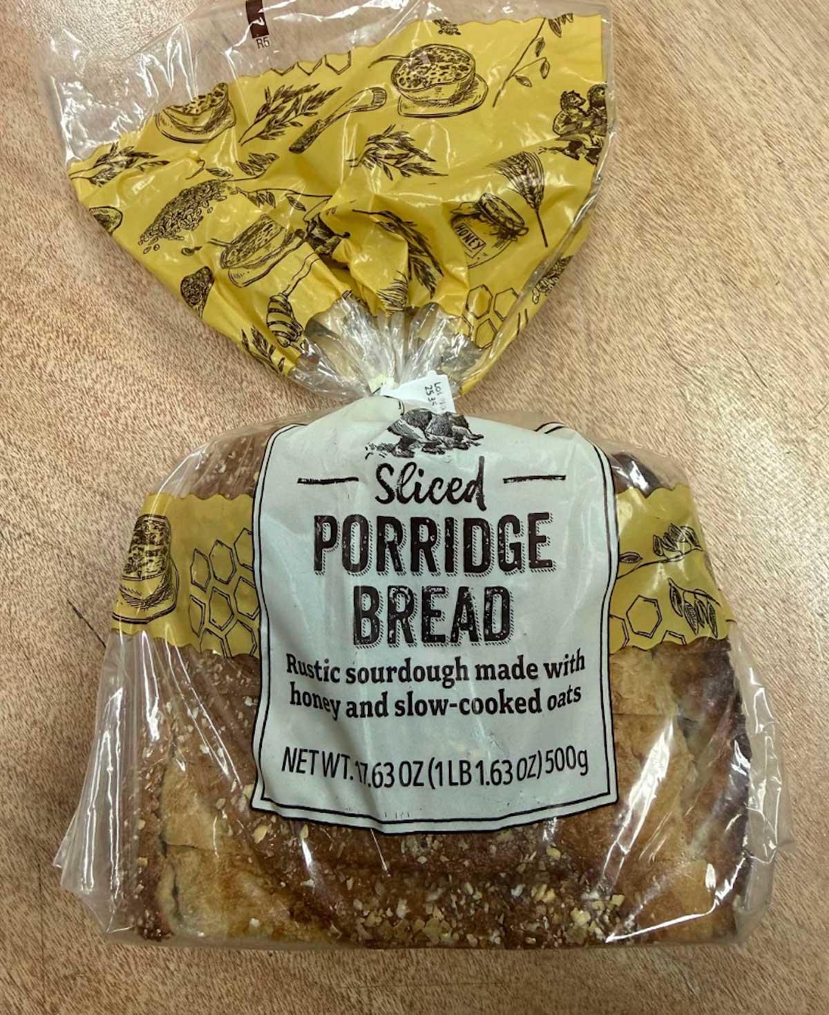 A loaf of sliced porridge bread in clear packaging, labeled "Rustic sourdough made with honey and slow-cooked oats." The label shows the net weight as 17.63 oz (500g). The package is on a wooden surface.
