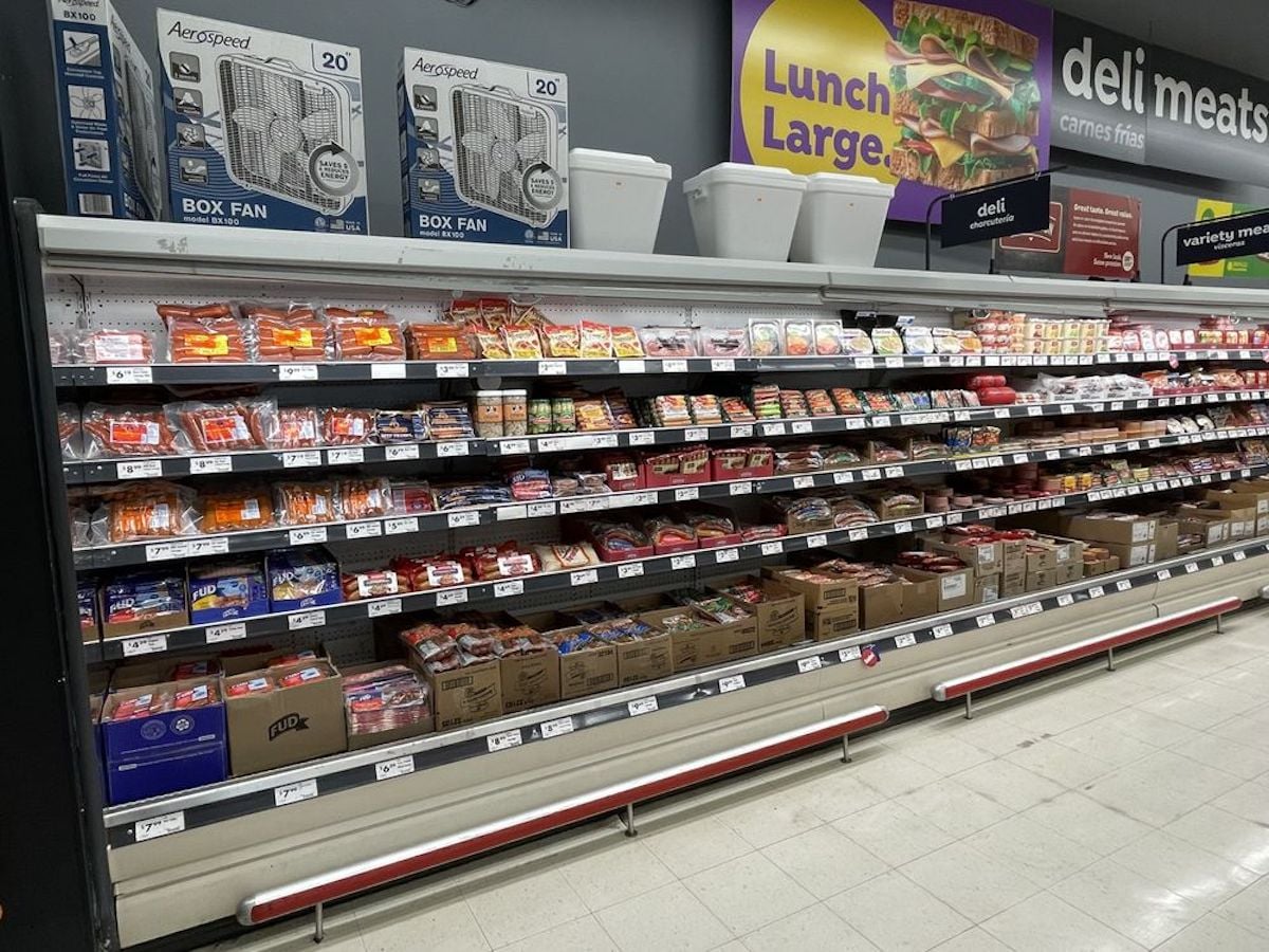 A grocery store deli meat section with multiple shelves filled with packaged meats. Box fans are stacked on top of the shelves, and a “Lunch Large” sign is displayed above. The floor is white and the area is well-lit.