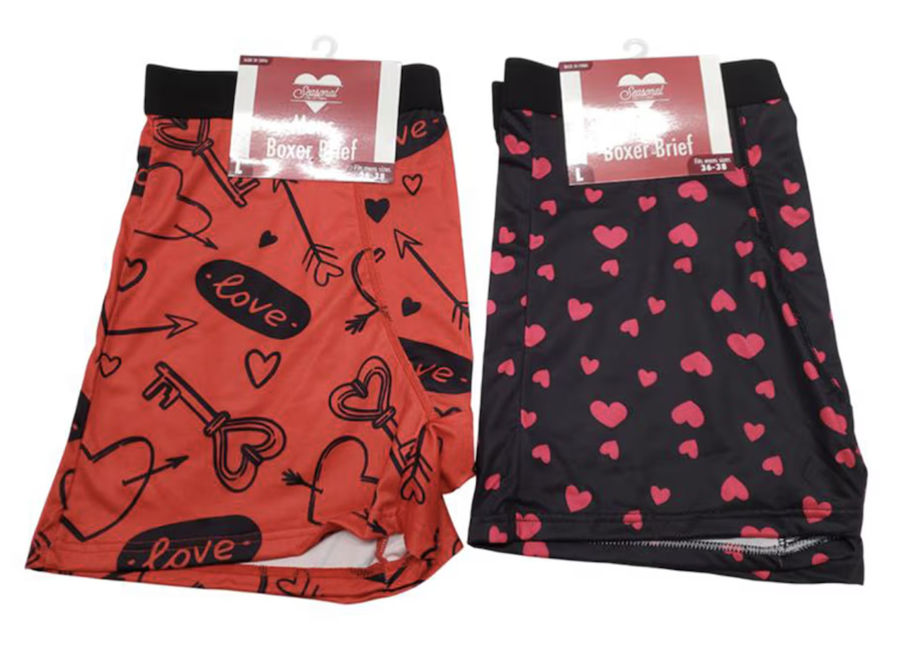 Two pairs of Valentine's Day-themed boxer briefs, one red with black love and arrow designs, and one black with pink heart patterns. Both have waistband tags labeled "Boxer Brief.