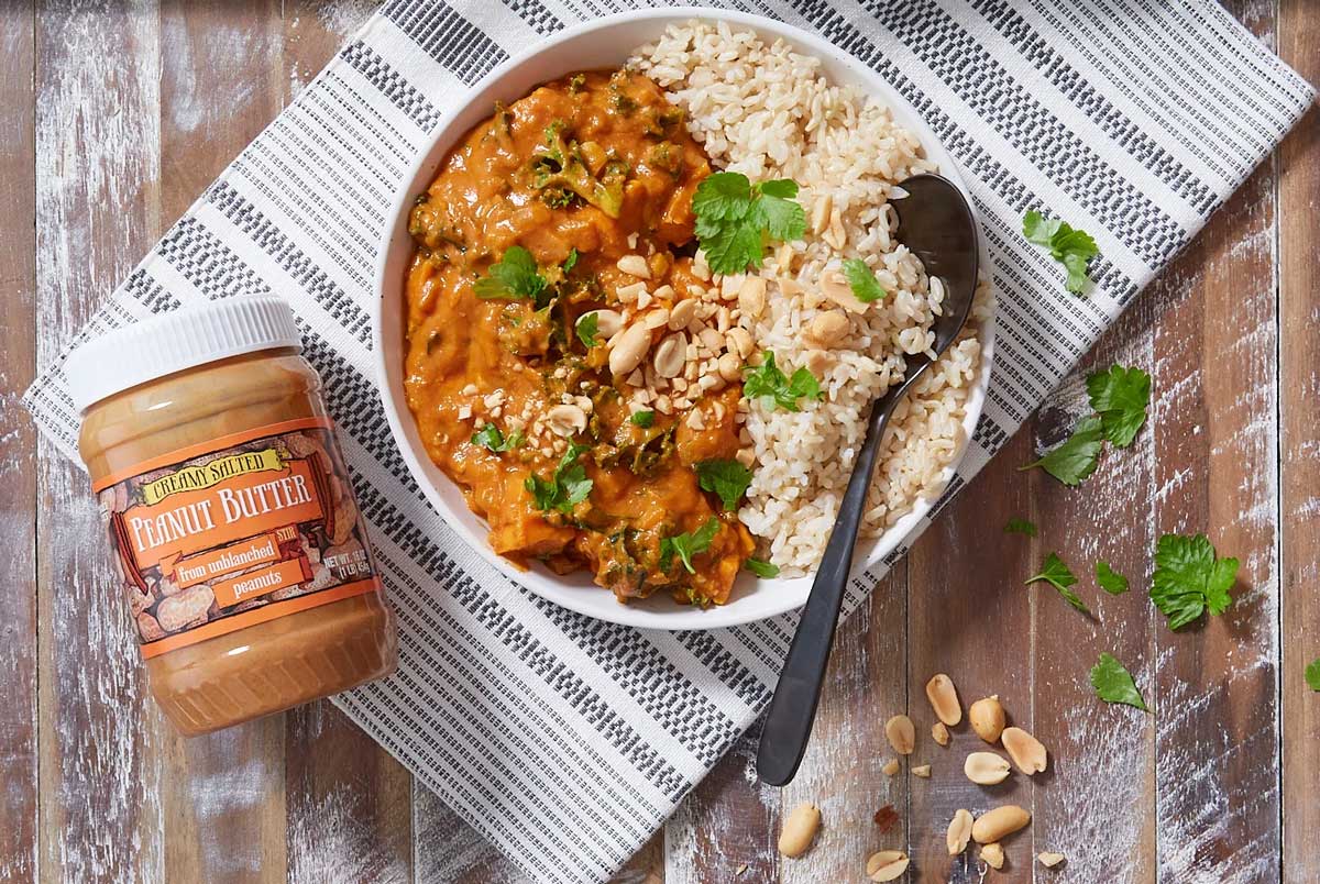 A bowl of rice and peanut stew garnished with cilantro and chopped peanuts sits on a striped cloth, with a spoon in the bowl. Nearby is a jar of creamy peanuts and salt peanut butter and scattered peanuts on a rustic wooden table.
