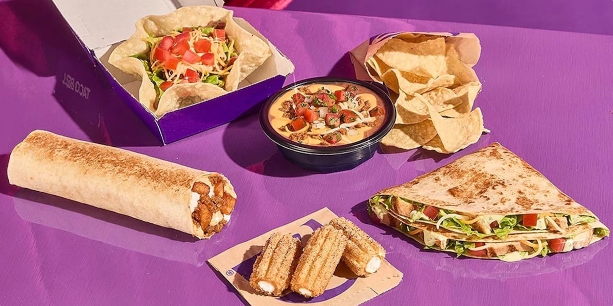 A variety of Tex-Mex fast food items on a purple surface, including a burrito, a taco salad, nachos with cheese dip, tortilla chips, a quesadilla, and two churros.