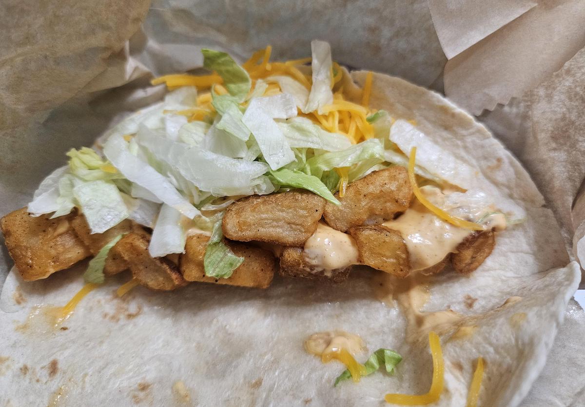 A soft tortilla filled with crispy fried potato wedges, shredded lettuce, shredded cheddar cheese, and a creamy orange sauce, partially wrapped in paper.