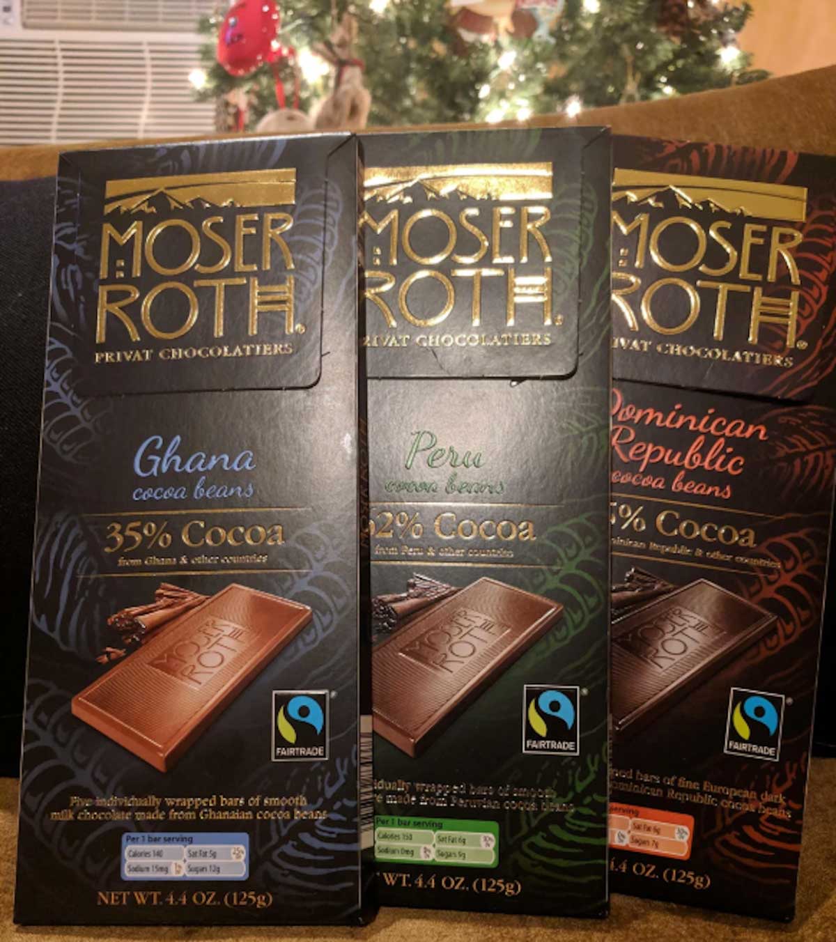 Three Aldi fan favorites—Moser Roth chocolate bars in Ghana 35% Cocoa, Peru 62% Cocoa, and Dominican Republic 70% Cocoa—are displayed upright in front of a glowing Christmas tree.