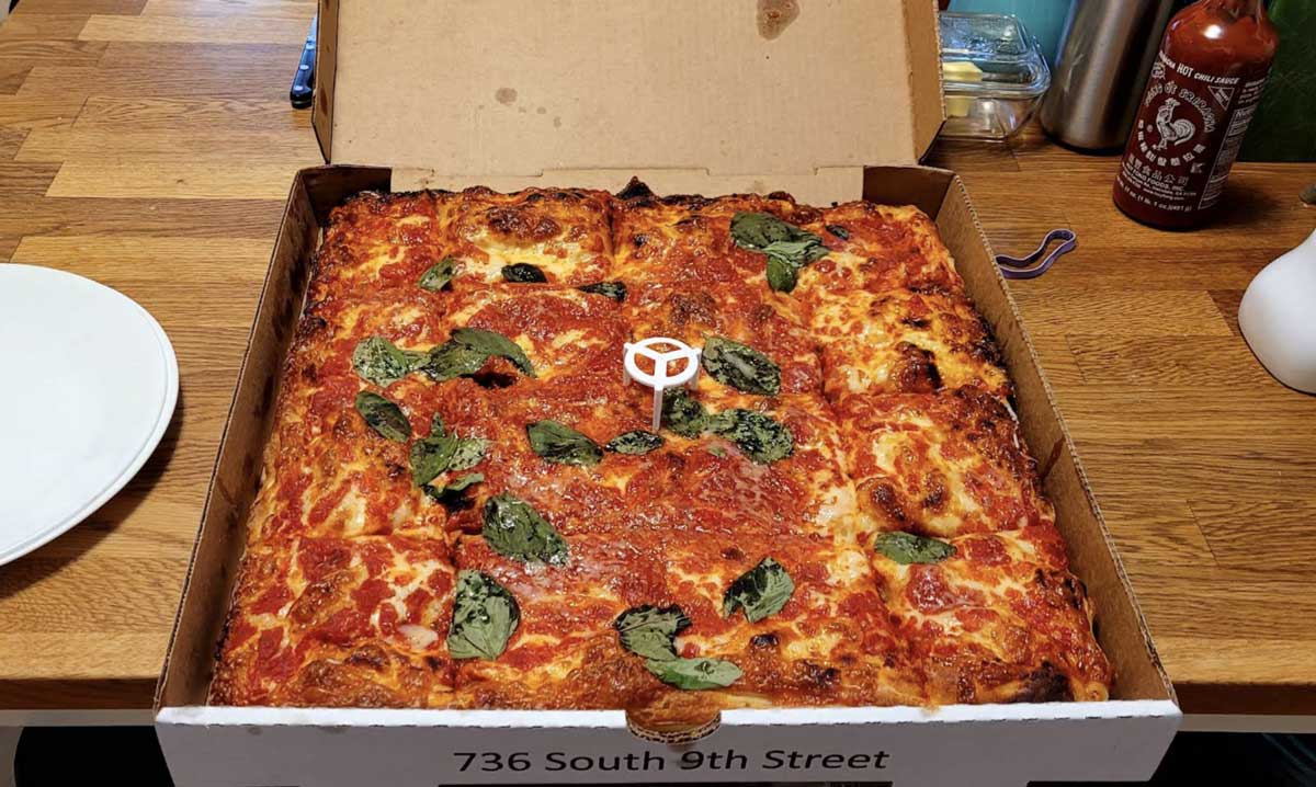 A square pizza reminiscent of classic East Coast cheap pizza slices, topped with tomato sauce, melted cheese, and fresh basil, sits in an open cardboard box on a wooden table. Bottles and a white plate are visible in the background.