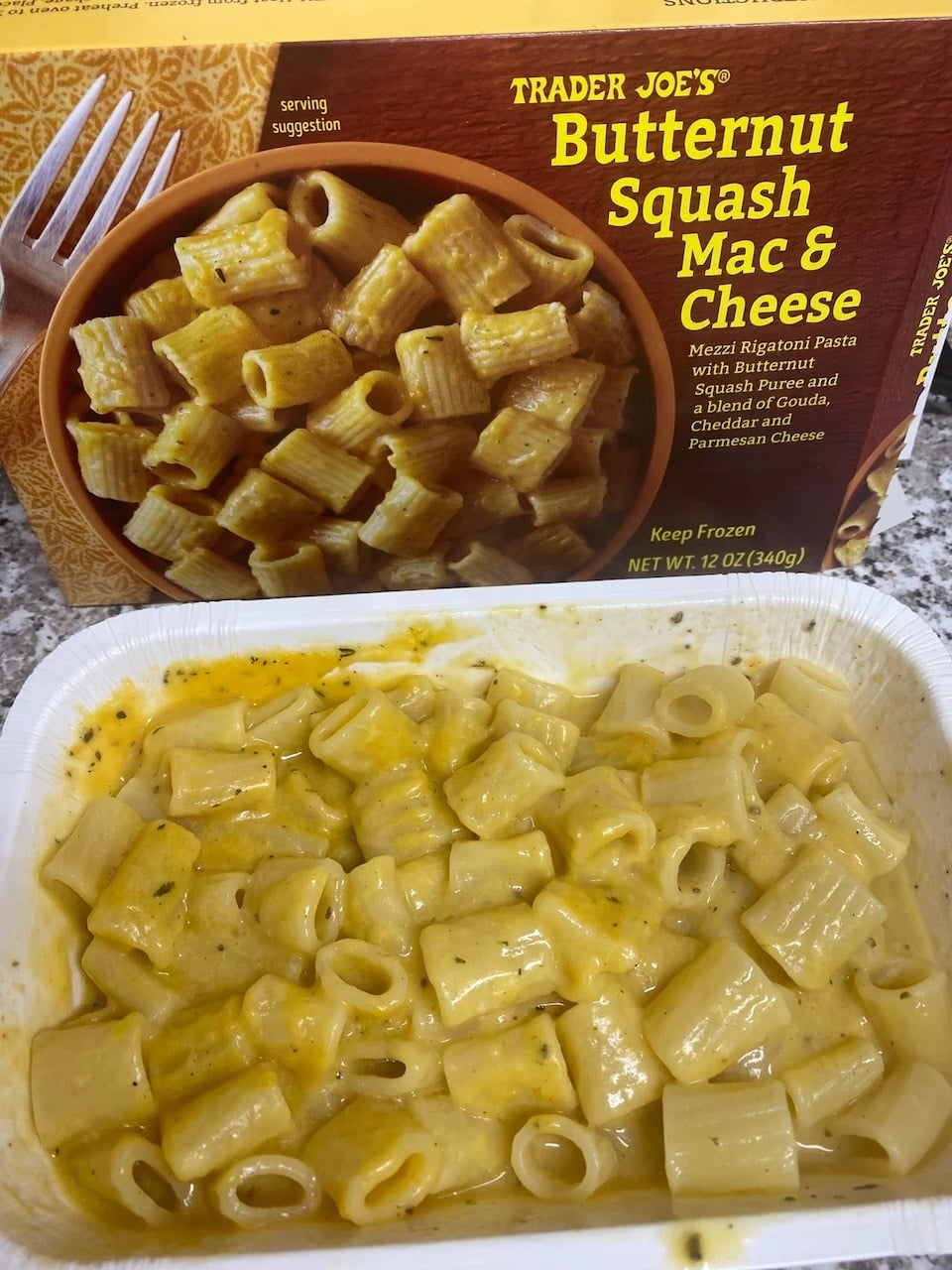A tray of Trader Joe’s Butternut Squash Mac & Cheese sits in front of its packaging, showing rigatoni pasta coated in a creamy yellow butternut squash cheese sauce. Trader Joe’s fall products.