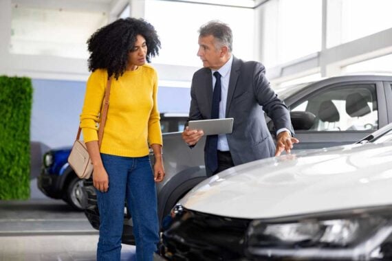 A car salesman in a suit shows a white car to a woman in a yellow sweater inside a showroom, using a tablet to explain details about the vehicle and car payments as she listens attentively.
