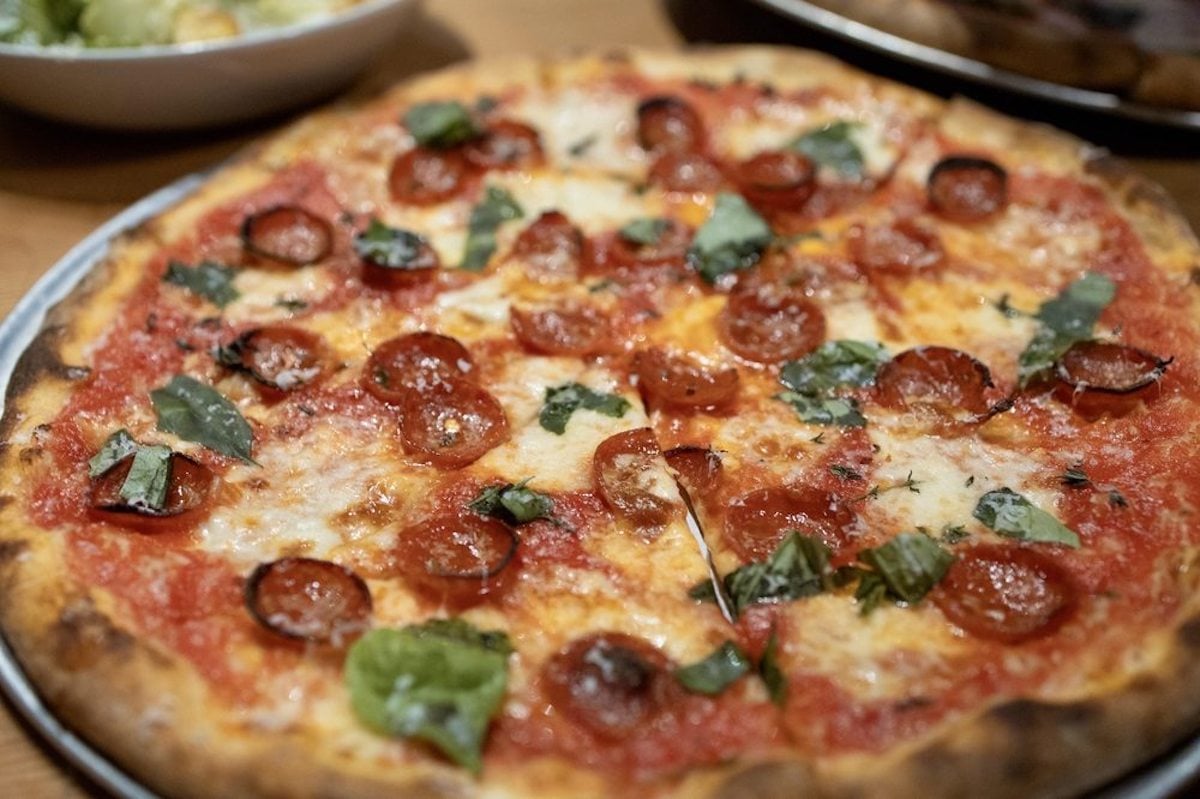 A close-up of a pepperoni pizza with melted cheese, tomato sauce, fresh basil leaves, and a thin crispy crust, served on a metal tray.