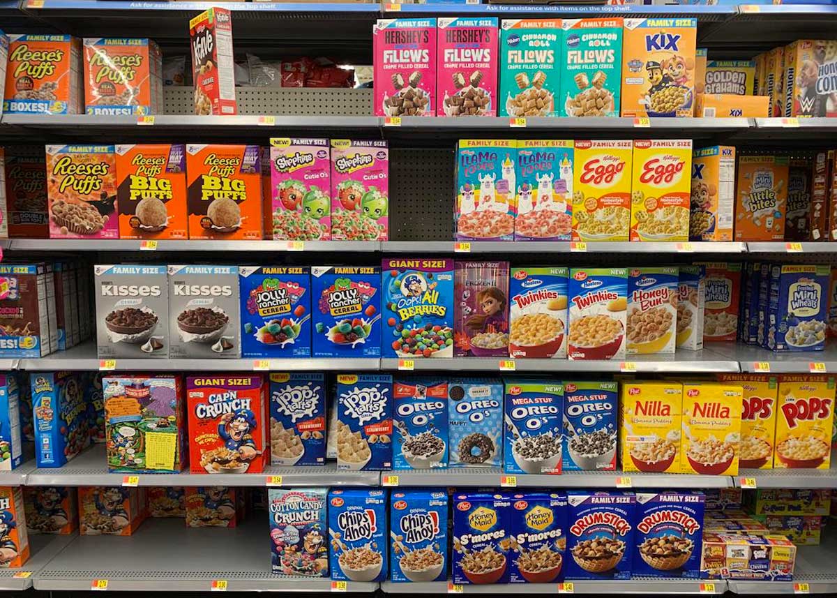 Grocery store shelves filled with a variety of colorful cereal boxes, including brands like Reese’s Puffs, Froot Loops, Lucky Charms, Cap’n Crunch, Oreo O’s, and more—some of the most expensive grocery items—stacked in multiple rows.