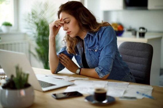 A woman sits at a desk with her hand on her forehead, looking stressed. She is surrounded by papers, a laptop, a phone, and a cup of coffee in a bright, modern home office setting.