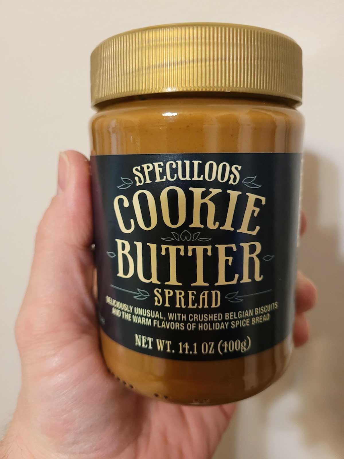 A hand holding a jar of Speculoos Cookie Butter Spread with a dark blue label. This overrated Trader Joe's favorite contains 14.1 ounces (400g) of spread made with crushed Belgian biscuits and holiday spice flavor.