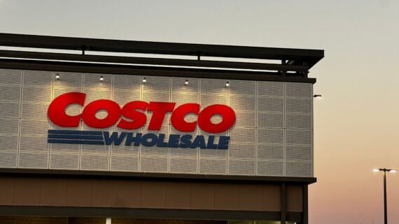 The exterior of a Costco Wholesale store at dusk, with the red and blue Costco Wholesale logo illuminated against a gray tiled wall. The sky in the background shows a gradient from light to dark.