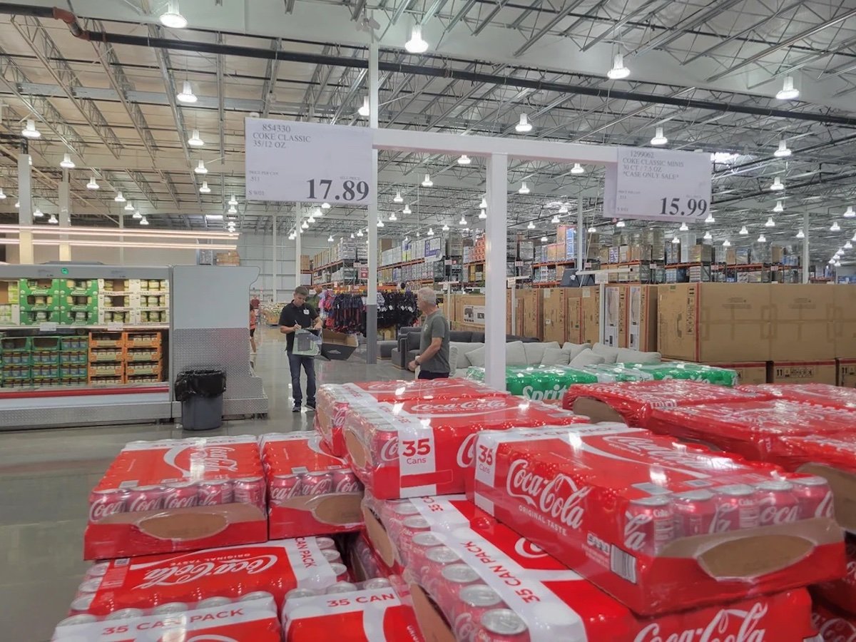 Stacks of Coca-Cola 35-can packs in a warehouse store, with shoppers in the background. Overhead signs display prices, and shelves with various products line the spacious, brightly lit interior.