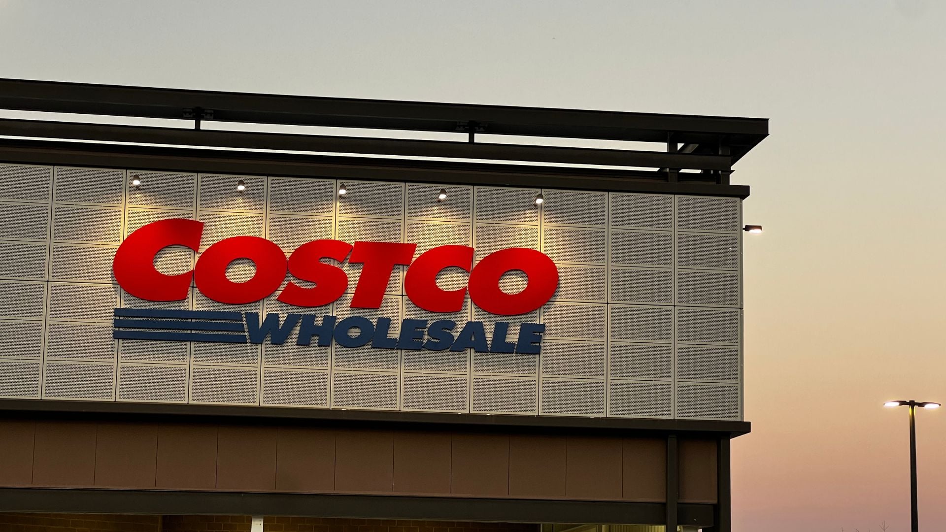 The exterior of a Costco Wholesale store at dusk, with the red and blue Costco Wholesale logo illuminated against a gray tiled wall. The sky in the background shows a gradient from light to dark.