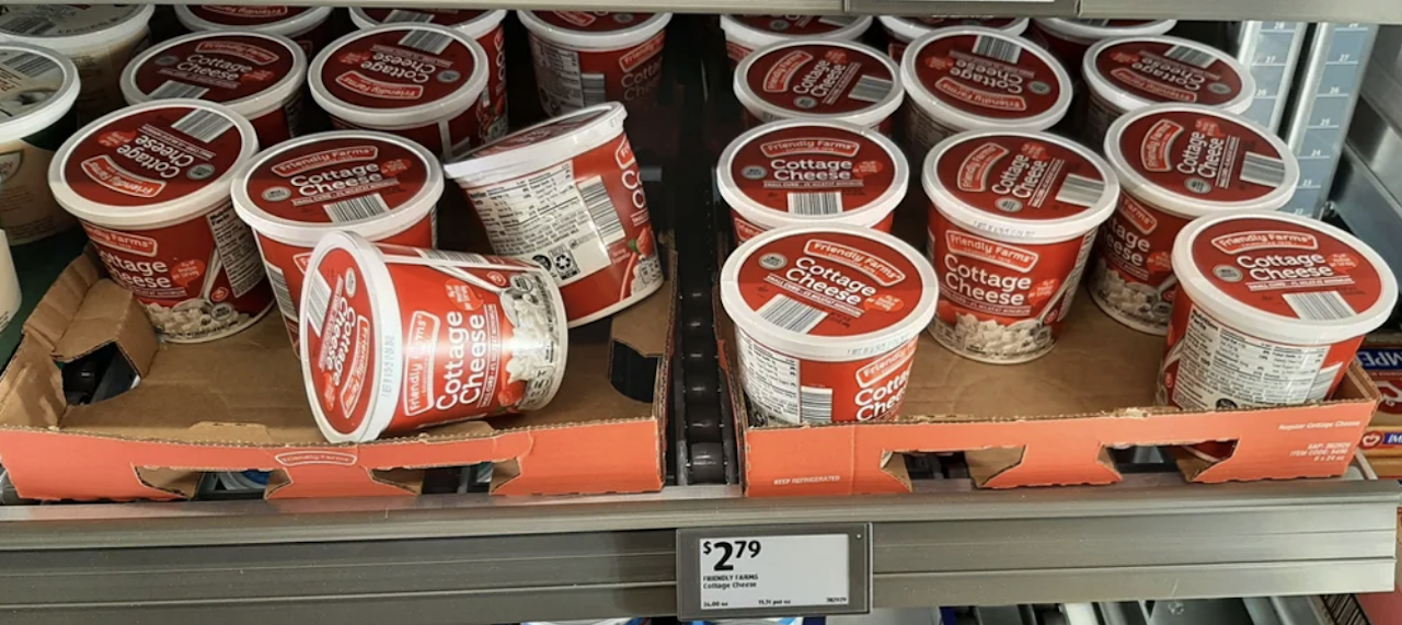 Several containers of cottage cheese with red and white labels are stacked on cardboard trays in a refrigerated grocery store shelf. A price tag below shows $2.79.