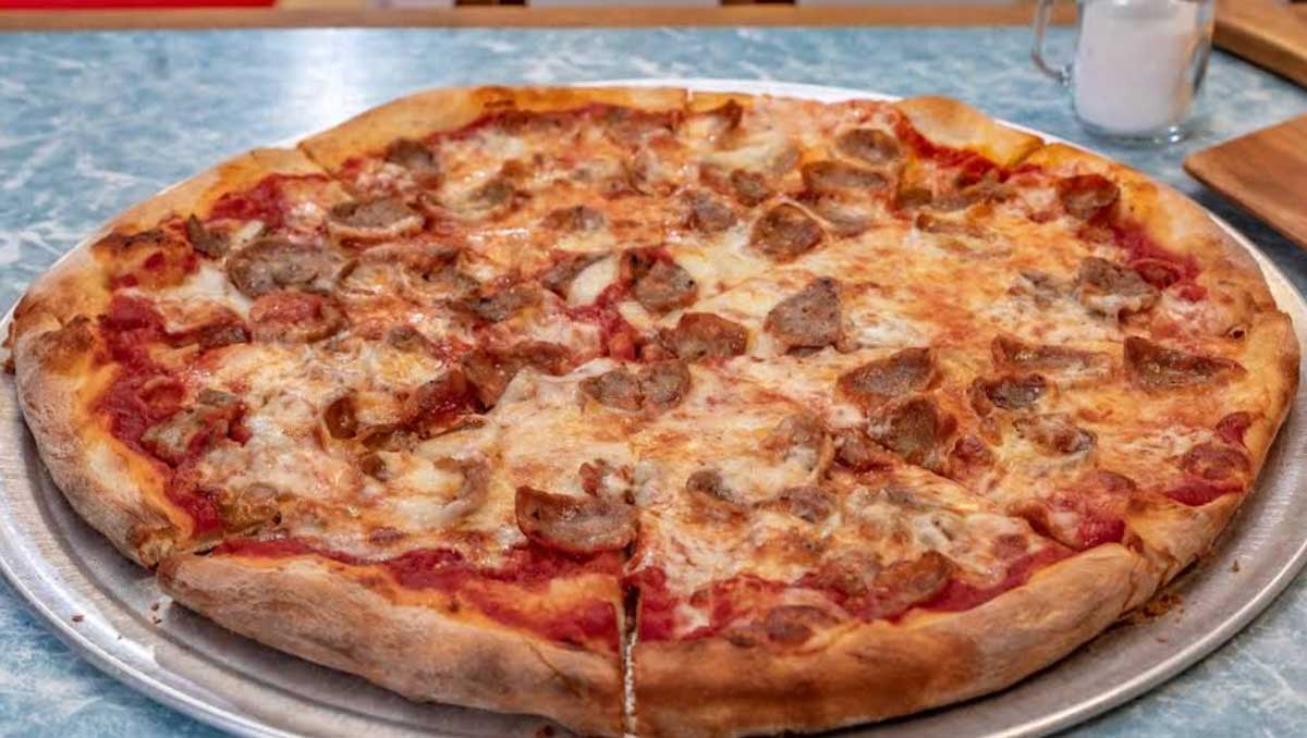 A large pizza topped with sausage and melted cheese sits on a metal tray on a blue marbled table, offering the classic flavor of East Coast cheap pizza slices, with a shaker of grated cheese or spices visible in the background.