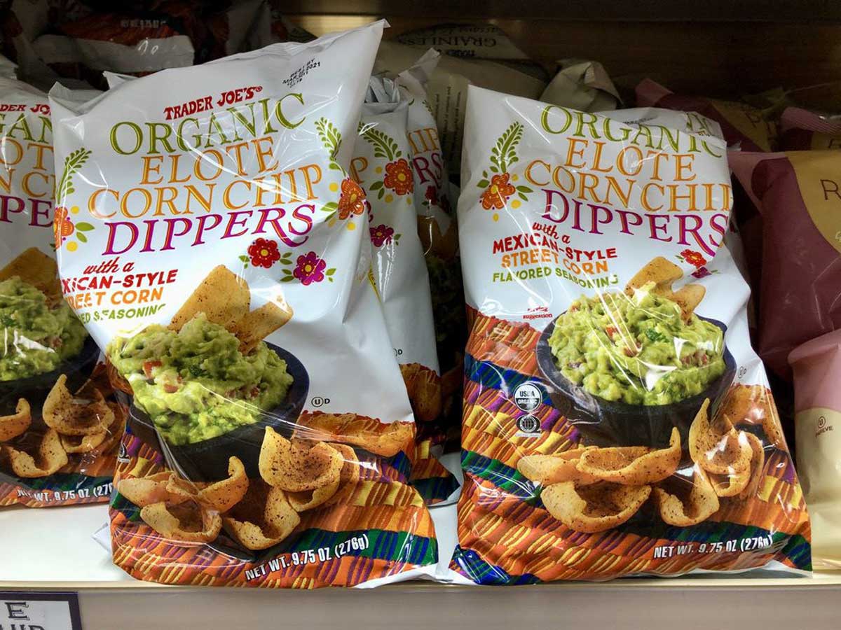 Bags of Trader Joe’s Organic Elote Corn Chip Dippers are displayed on a shelf. The packaging shows corn chips with guacamole and mentions Mexican-style street corn flavoring and seasoning.