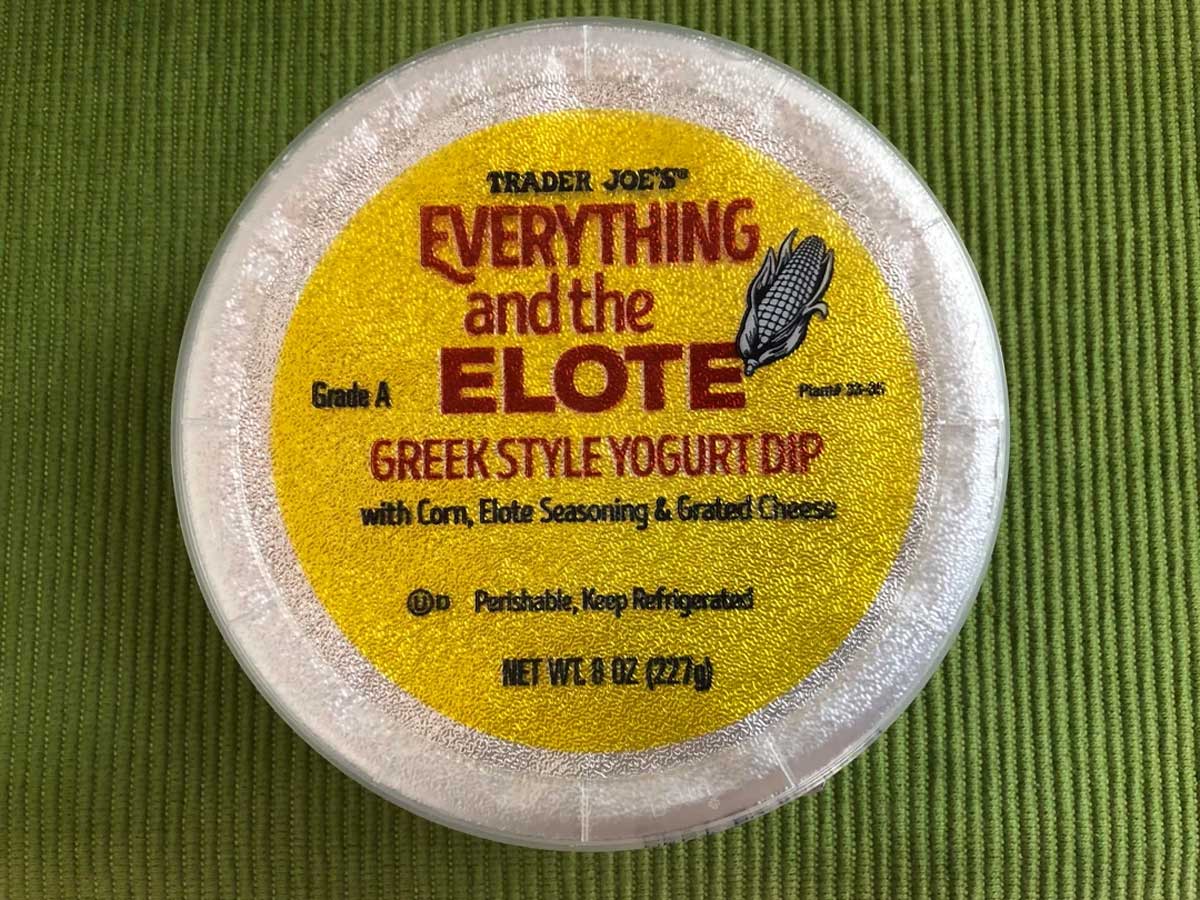 A round container of Trader Joe’s Everything but the Elote Greek Style Yogurt Dip, featuring a yellow label with red and brown text and a corn graphic, placed on a green textured surface.