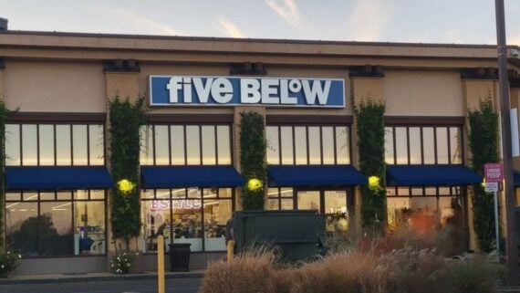 A Five Below store with large windows, blue awnings, and greenery climbing the building exterior. The store&rsquo;s sign is prominently displayed above the entrance. A dumpster and parking lot are visible in front.