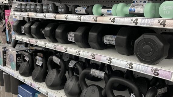 Shelves stocked with various weights of dumbbells and kettlebells in a store, each labeled with prices ranging from $4 to $5. The weights are neatly arranged in rows on white shelves.