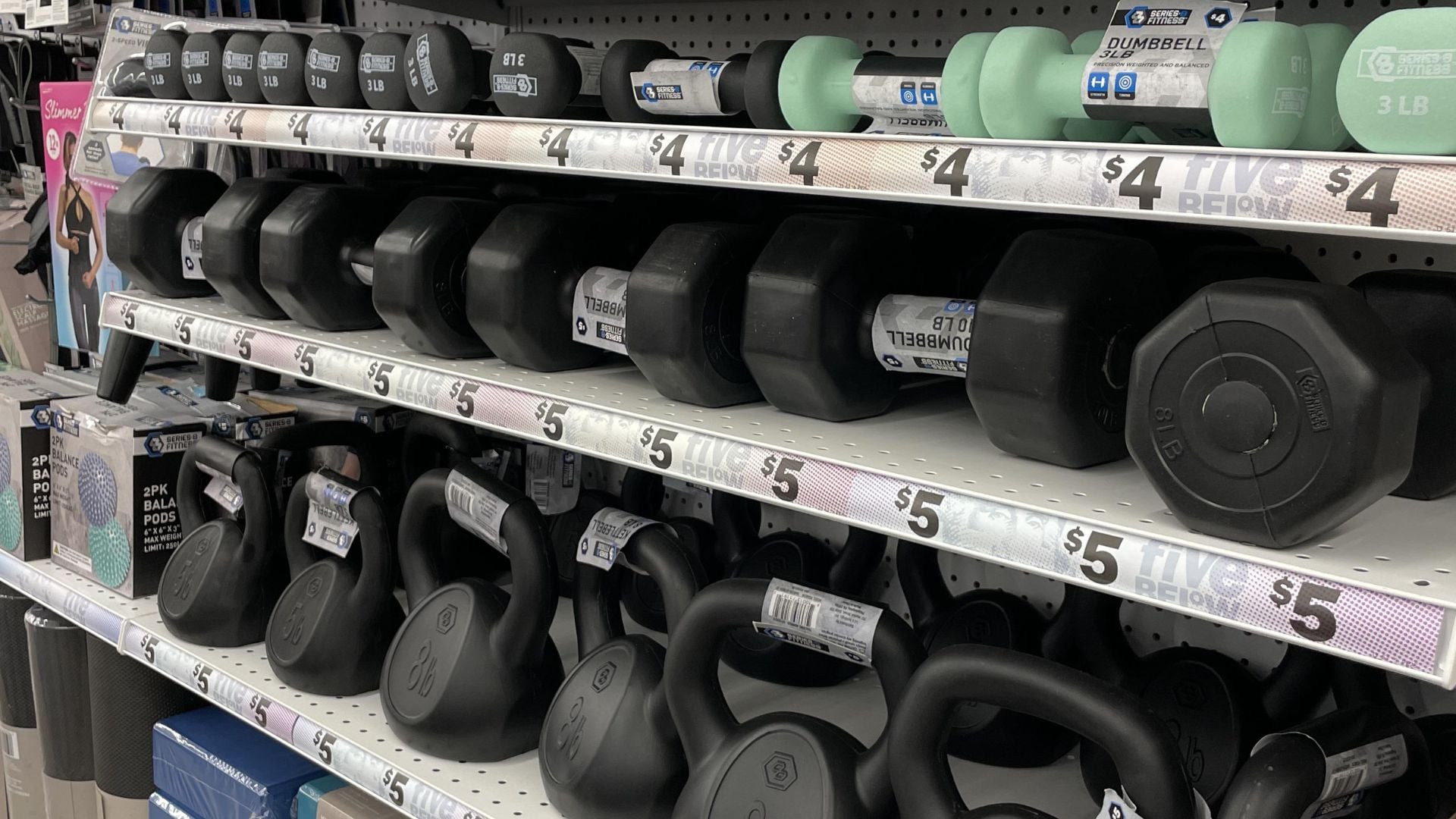 Shelves stocked with various weights of dumbbells and kettlebells in a store, each labeled with prices ranging from $4 to $5. The weights are neatly arranged in rows on white shelves.
