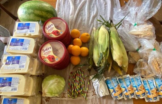 A table at the food pantry is filled with blocks of Swiss cheese, tubs of hummus, a watermelon, ears of corn, oranges, lettuce, packs of string cheese, colorful candy ropes, and bags of bread.