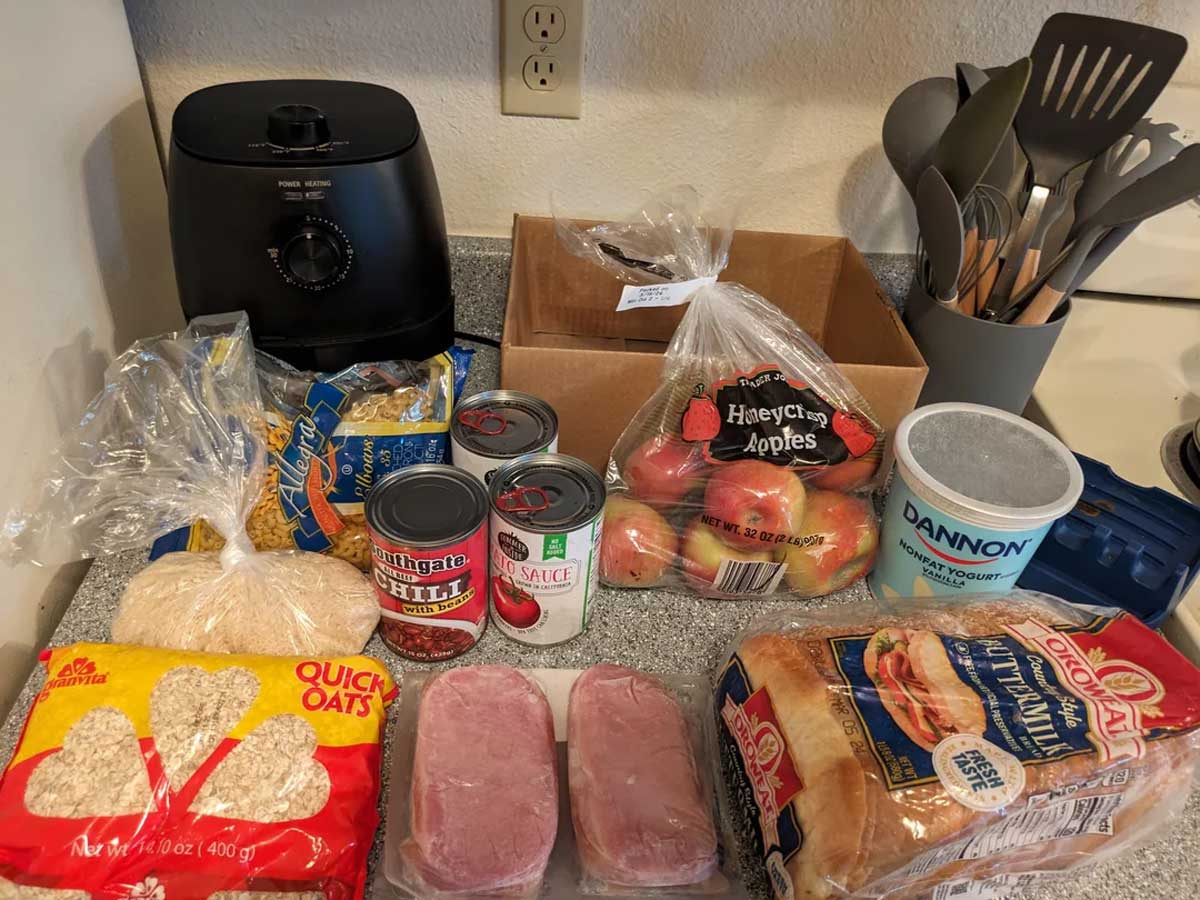 A kitchen counter displays an air fryer, utensils, bread, quick oats, apples, canned chili, tomato sauce, vanilla yogurt, and two packages of uncooked pork chops.