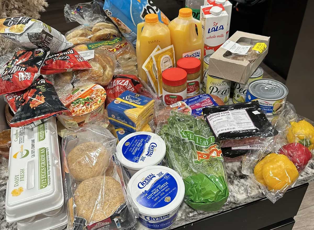 A variety of groceries on a kitchen counter, including eggs, bread, bagels, orange juice, cheese, vegetables, instant noodles, chips, milk, canned goods, cream cheese, and seasoning packets.