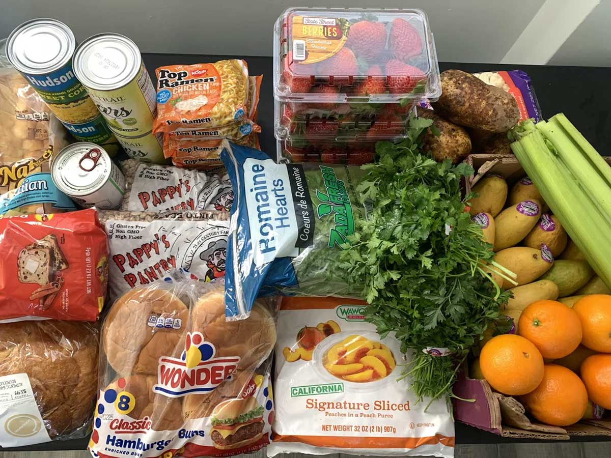 A variety of groceries on a table, including bread, hamburger buns, ramen noodles, canned food, strawberries, bananas, oranges, celery, romaine lettuce, cilantro, pears, and sliced cheese.