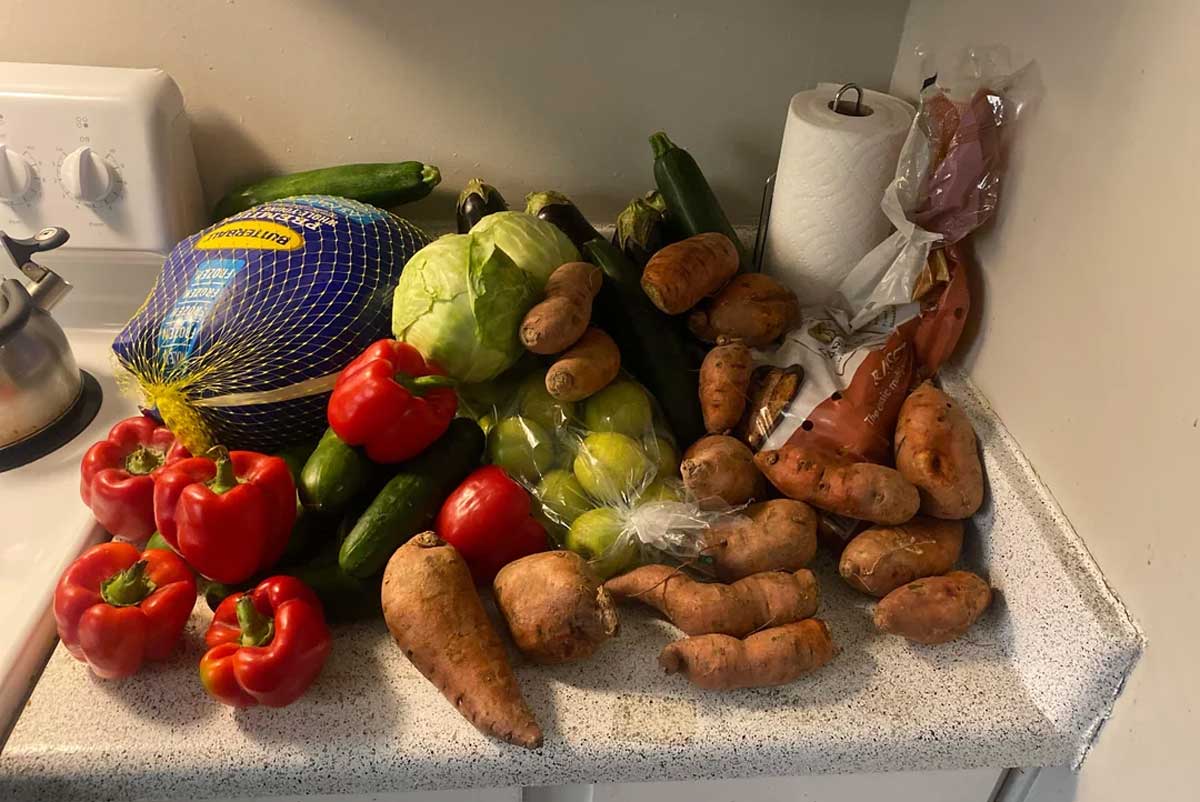 A kitchen counter with various vegetables and a turkey. Visible items include red bell peppers, sweet potatoes, eggplants, zucchinis, a cabbage, cucumbers, limes, a bagged turkey, paper towels, and a kettle on the stove.