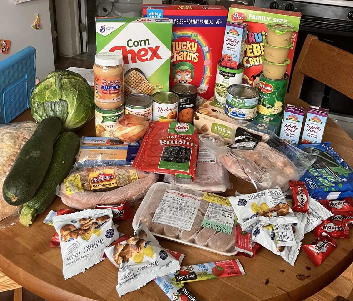 A wooden table covered with various groceries, including cereal boxes, fresh vegetables, canned goods, packaged meats, applesauce, cream cheese, snack bars, and frozen foods.