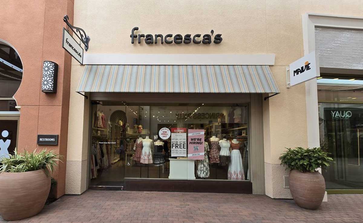 Storefront of Francesca’s boutique with striped awning, mannequins in dresses on display, and a francesca's closing sale sign in the window. Large potted plants flank the entrance. Signs for nearby stores are visible.