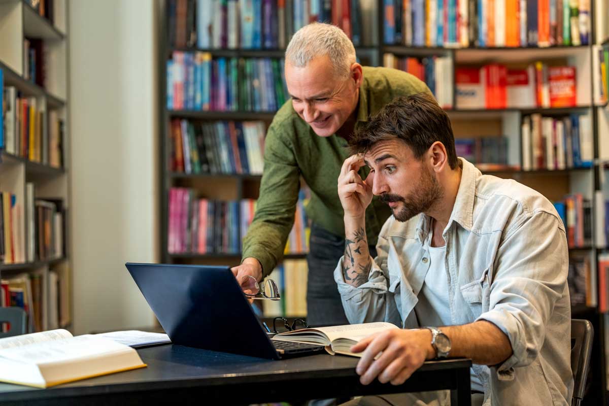 Two men in a library, one older and standing, the other younger and seated at a table with a laptop and open books. The standing man smiles and points at the laptop while the younger man looks focused.