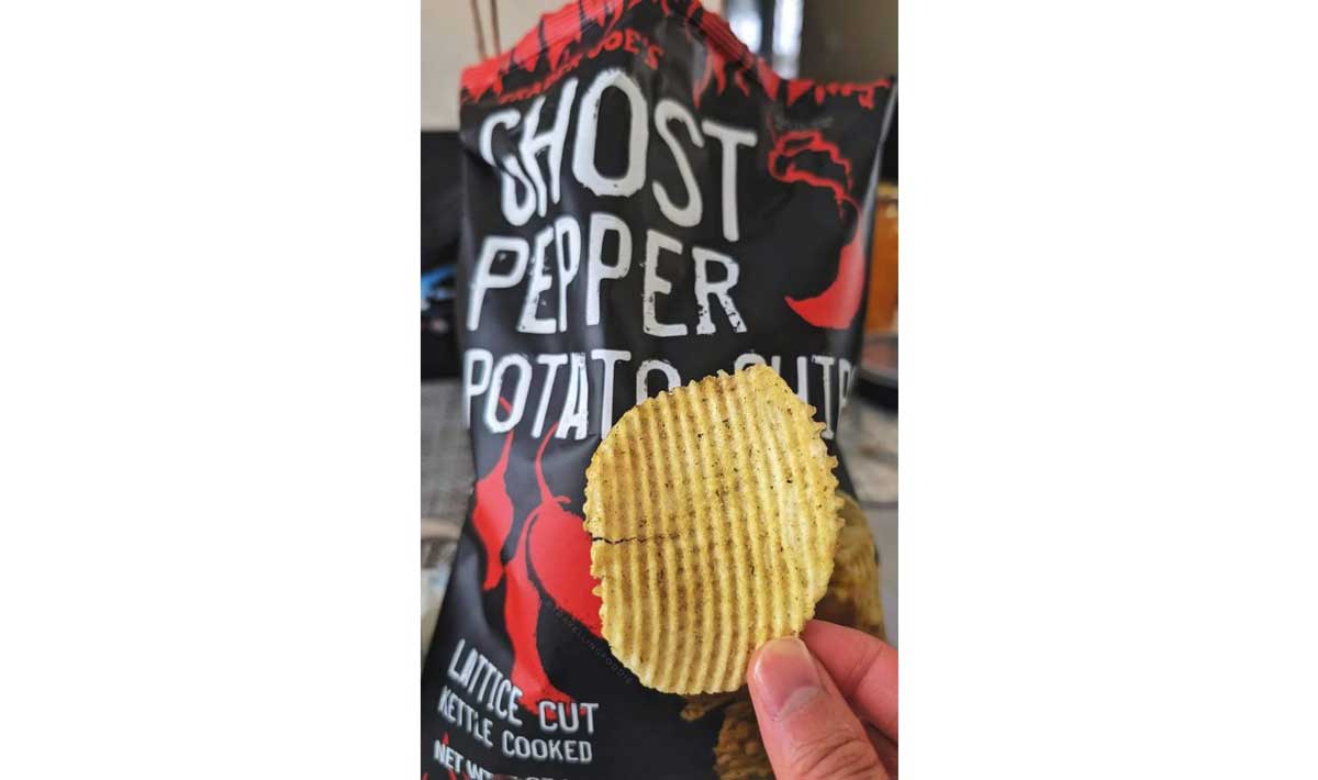 A hand holds a ridged potato chip in front of an open bag labeled "Ghost Pepper Potato Chips" with bold white letters and red accents.