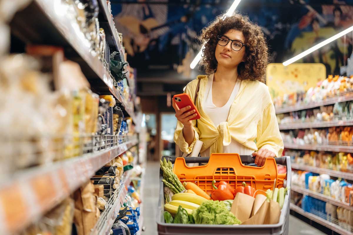 A person with curly hair and glasses pushes a shopping cart filled with groceries, including vegetables and bananas, down an aisle in a supermarket while holding a red smartphone.