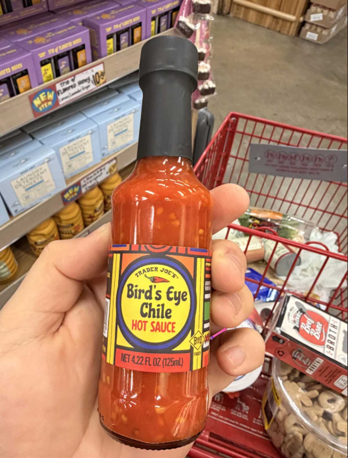 A hand holds a bottle of Trader Joe’s Bird’s Eye Chile Hot Sauce in a grocery store, with shelves and a red shopping cart containing various items visible in the background.