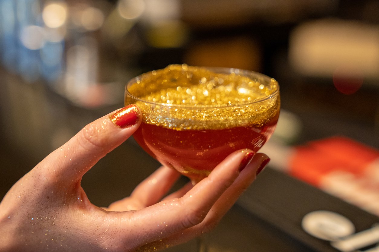 A hand with red-painted nails holds a cocktail glass filled with a red drink, sparkling with gold glitter. The rim of the glass and the hand are also dusted with glitter.