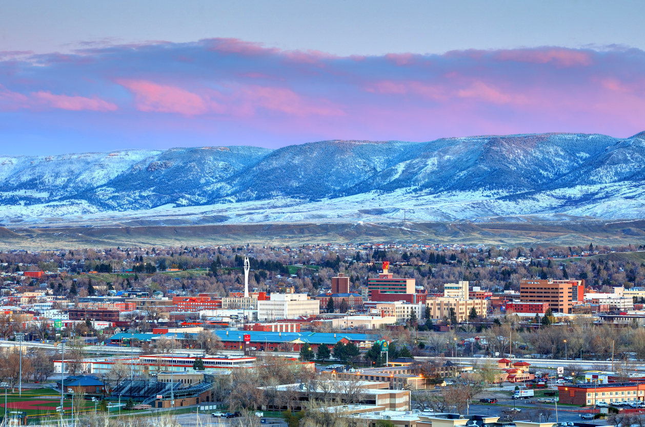 Cityscape of Casper, Wyoming at sunset, with buildings in the foreground, sparse trees, and snow-covered mountains in the background under a pink and blue sky.