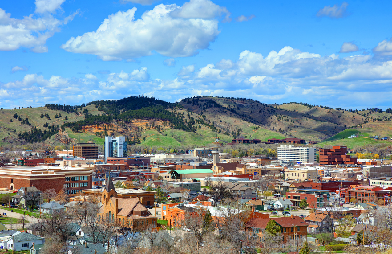 A cityscape with a mix of modern and historic buildings set against green, rolling hills and a partly cloudy blue sky in the background.