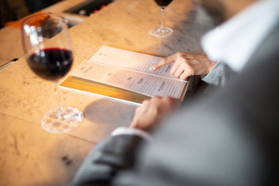 A person in a suit sits at a marble counter, pointing at a menu while holding it open. Two glasses of red wine are on the counter, creating a relaxed, upscale dining atmosphere.