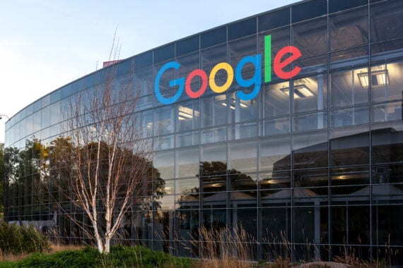 A modern glass building with the colorful Google logo displayed on the exterior. Leafless trees and greenery are visible in the foreground, with reflections of the sky and surroundings on the windows.