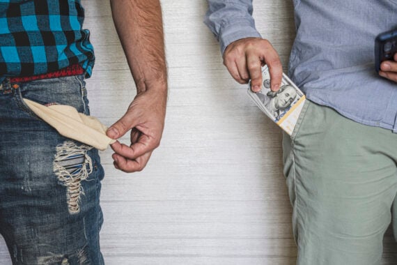 Two men standing side by side; one with an empty, pulled-out pocket and distressed jeans, the other wearing green pants, holding a stack of U.S. hundred-dollar bills, highlighting a contrast in financial situations.