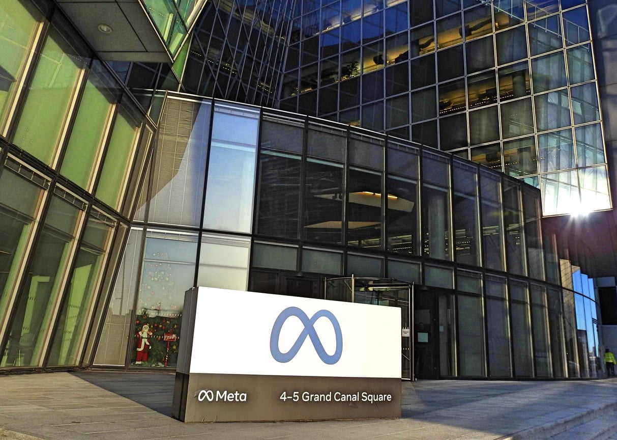 Exterior view of a modern glass office building with a Meta sign and logo displayed in front, showing the address "4-5 Grand Canal Square." Sunlight reflects off the building's surface.