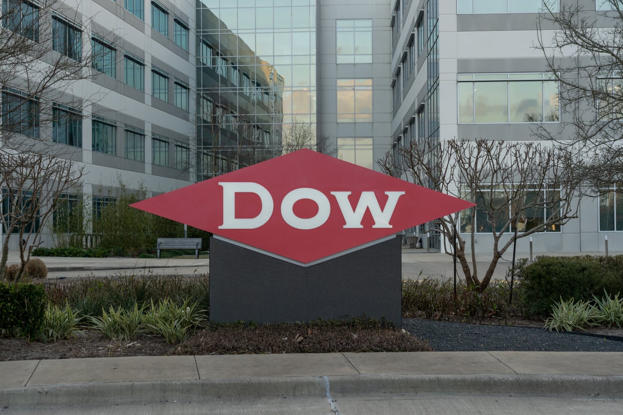 A large red diamond-shaped Dow sign sits in front of an office building, surrounded by low plants and leafless trees, reflecting uncertainty as the company faces 2026 layoffs.