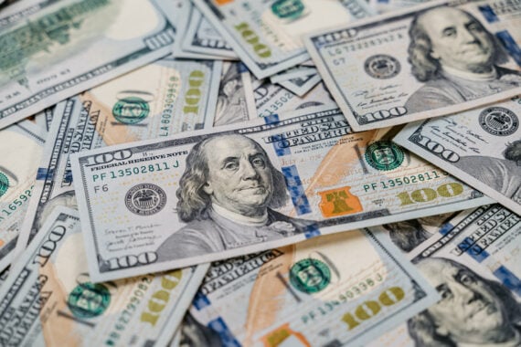 A pile of overlapping U.S. 100 dollar bills, featuring Benjamin Franklin&rsquo;s portrait, fills the frame. The bills show security features like blue security strips and orange ink.