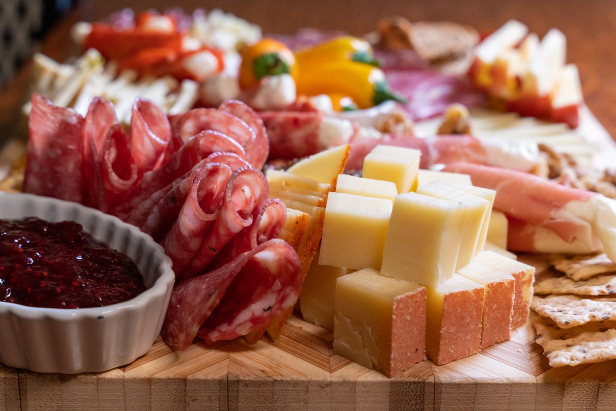 A close-up of a charcuterie board with sliced cured meats, stacked cheese cubes, crackers, fresh fruit, and a small dish of red jam on a wooden surface.