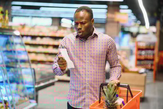 A man in a plaid shirt stands in a grocery store aisle, holding a shopping basket with a pineapple and looking at a receipt or shopping list with a surprised expression.