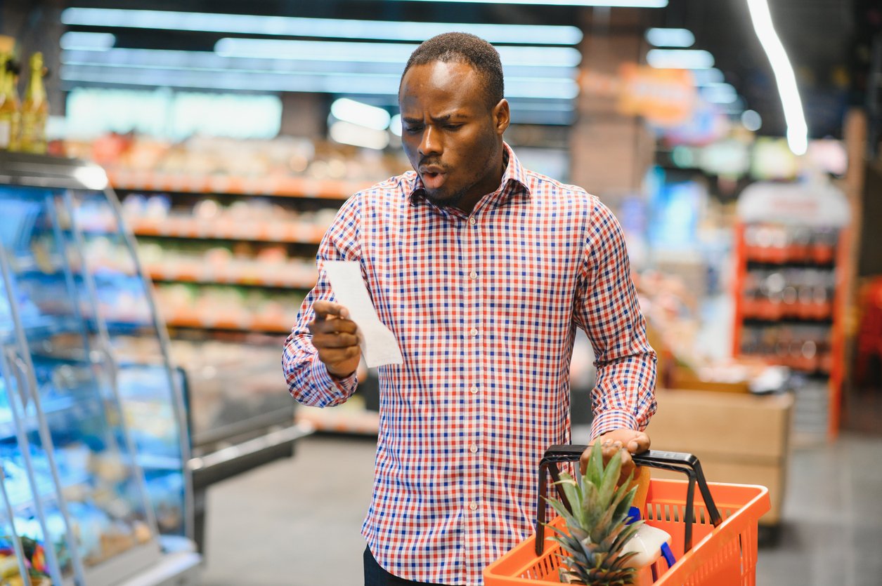 A man in a plaid shirt stands in a grocery store aisle, holding a shopping basket with a pineapple and looking at a receipt or shopping list with a surprised expression.