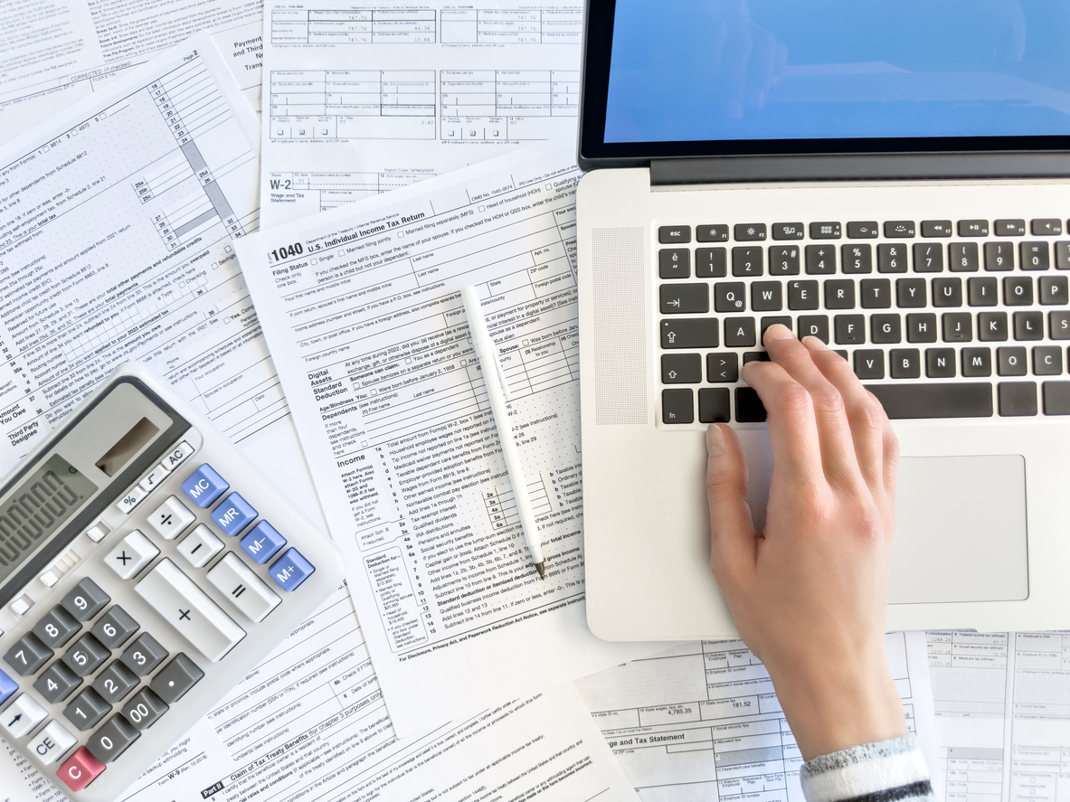 A person using a laptop surrounded by tax forms, a pen, and a calculator, suggesting they are working on taxes or financial documents.