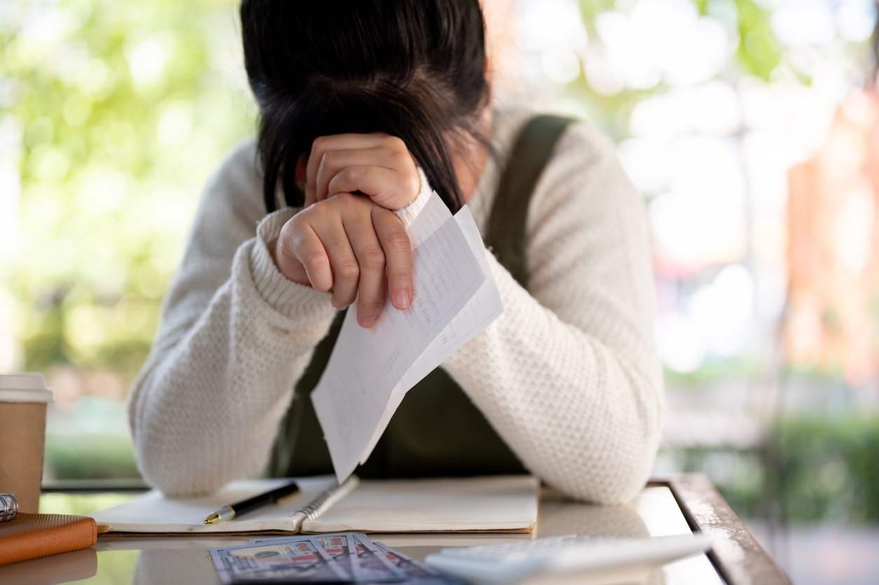 A person sits at a table with their head in their hands, holding a bill. Papers, a notebook, pen, cup, and money are on the table, suggesting financial stress or worry. Bright, blurred background with greenery.