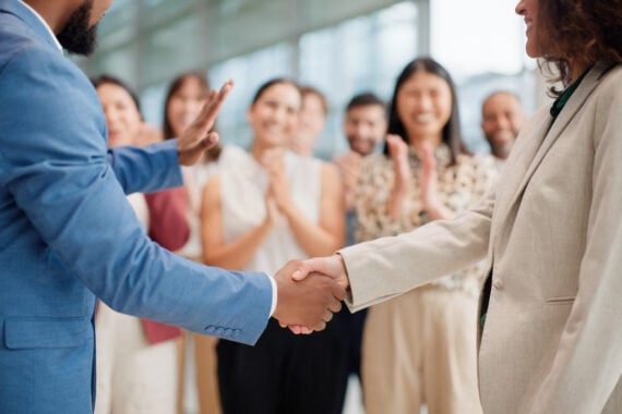 Two people in business attire shake hands while a group of colleagues stand in the background, smiling and clapping in a modern office setting.